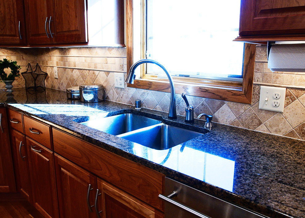 Example of a large classic l-shaped light wood floor eat-in kitchen design in Milwaukee with an undermount sink, raised-panel cabinets, medium tone wood cabinets, granite countertops, beige backsplash, stone tile backsplash, stainless steel appliances and an island