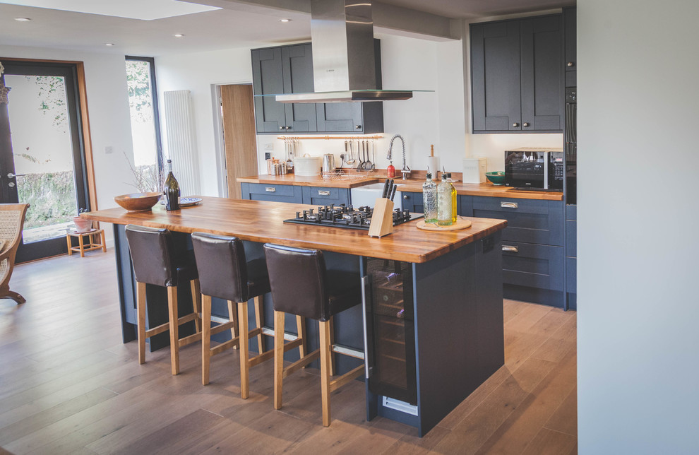 Design ideas for a medium sized rustic single-wall open plan kitchen in Devon with a belfast sink, shaker cabinets, grey cabinets, wood worktops, black appliances, laminate floors and a breakfast bar.
