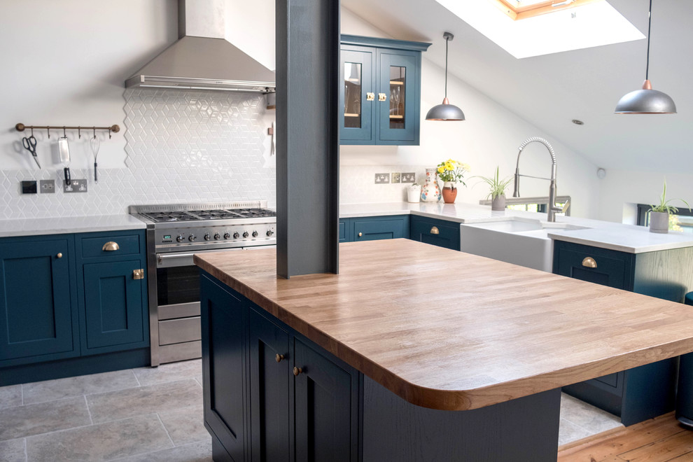 Medium sized country u-shaped open plan kitchen in London with a belfast sink, shaker cabinets, blue cabinets, white splashback, porcelain splashback, stainless steel appliances, porcelain flooring, an island, wood worktops and grey floors.