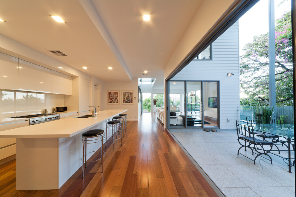 This is an example of a large contemporary galley kitchen/diner in Brisbane with a double-bowl sink, flat-panel cabinets, white cabinets, engineered stone countertops, white splashback, glass sheet splashback, stainless steel appliances, medium hardwood flooring and an island.