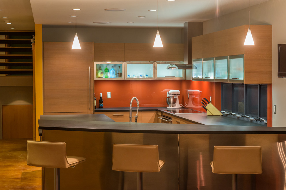 Modern inredning av ett mellanstort kök, med släta luckor, skåp i ljust trä, orange stänkskydd, glaspanel som stänkskydd, integrerade vitvaror, bänkskiva i täljsten, betonggolv, en halv köksö och brunt golv