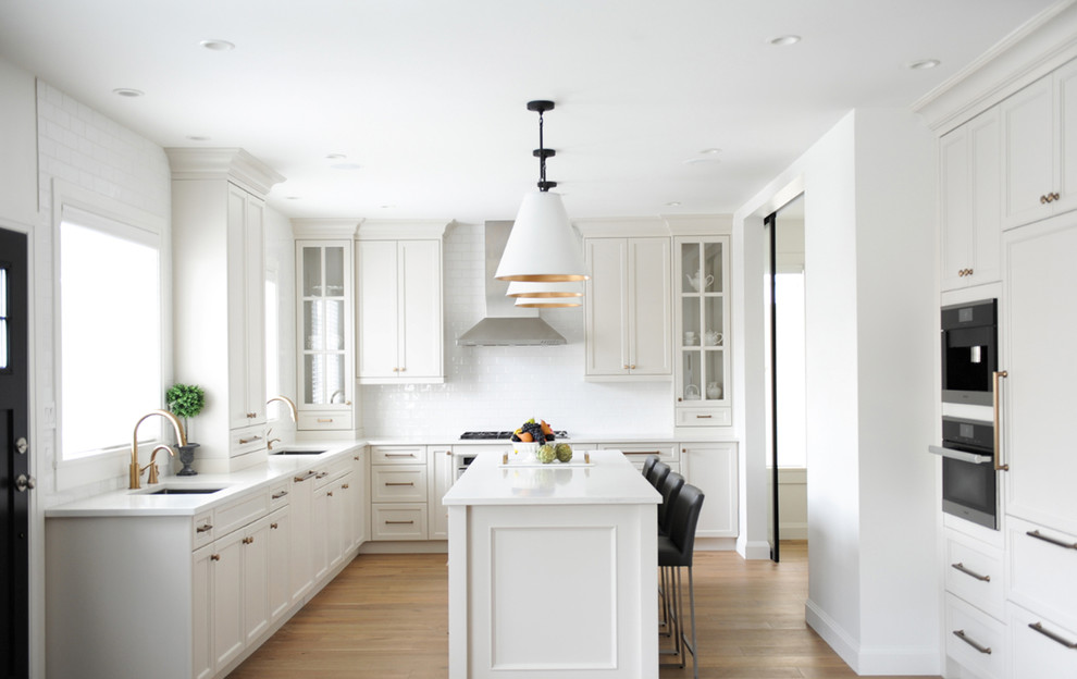 This is an example of a traditional l-shaped kitchen in Vancouver with white cabinets, a submerged sink, recessed-panel cabinets, white splashback, metro tiled splashback, light hardwood flooring, an island and beige floors.