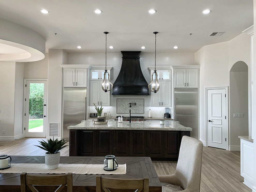 Example of a large transitional l-shaped porcelain tile, gray floor and vaulted ceiling open concept kitchen design in Phoenix with a farmhouse sink, raised-panel cabinets, white cabinets, quartzite countertops, white backsplash, ceramic backsplash, stainless steel appliances, an island and beige countertops