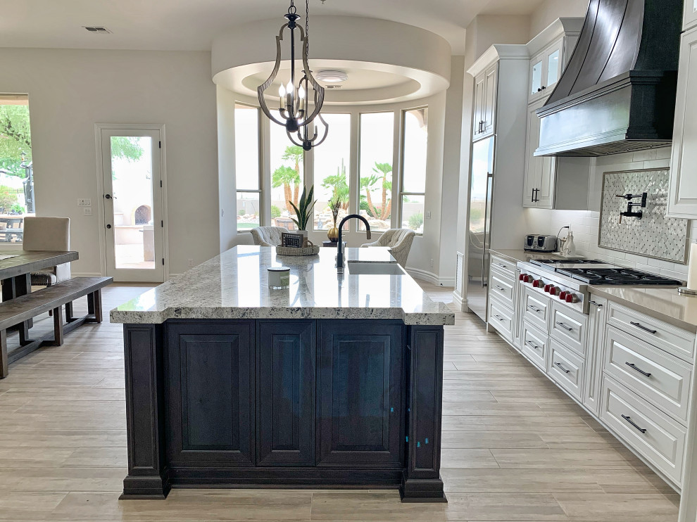 Large transitional l-shaped porcelain tile, gray floor and vaulted ceiling open concept kitchen photo in Phoenix with a farmhouse sink, raised-panel cabinets, white cabinets, quartzite countertops, white backsplash, ceramic backsplash, stainless steel appliances, an island and beige countertops