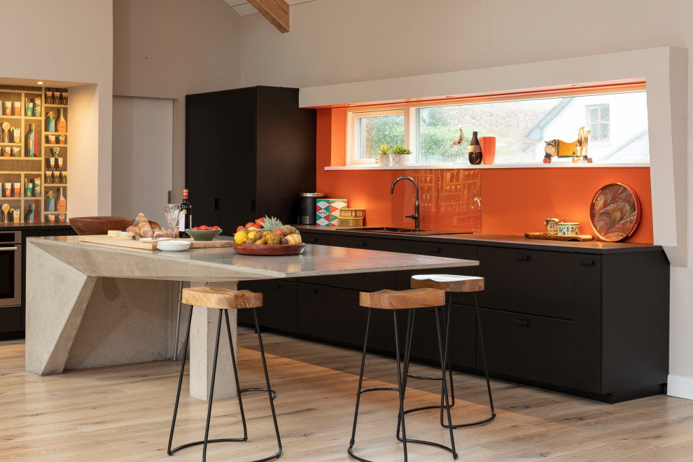 Idéer för mellanstora lantliga grått kök, med en nedsänkt diskho, släta luckor, svarta skåp, bänkskiva i betong, orange stänkskydd, integrerade vitvaror, ljust trägolv och en köksö