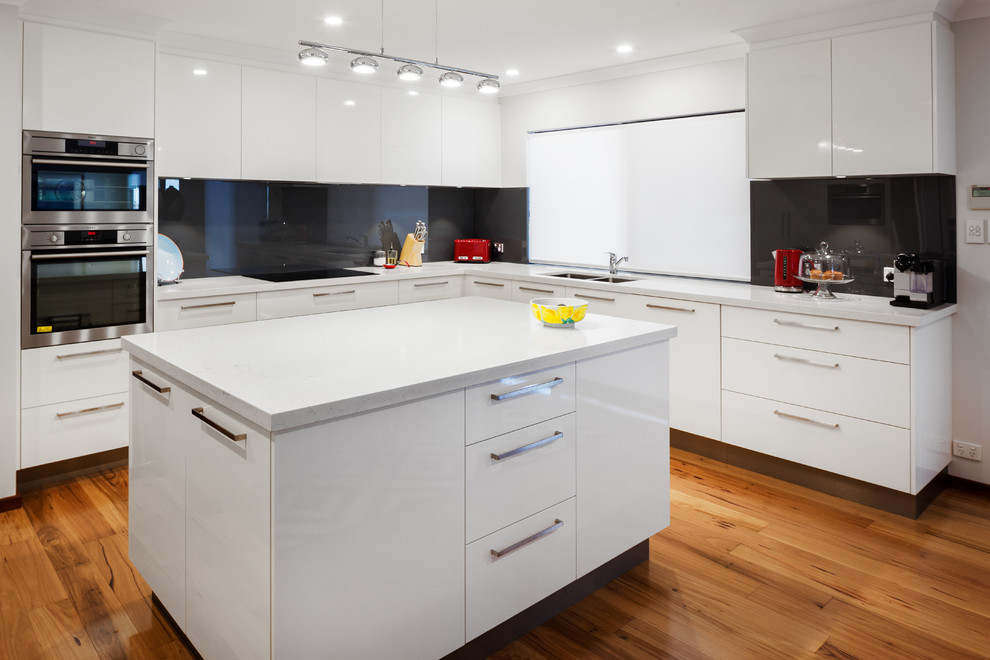 Bild på ett funkis vit vitt kök, med släta luckor, vita skåp, bänkskiva i kvarts, svart stänkskydd, glaspanel som stänkskydd, rostfria vitvaror och en köksö