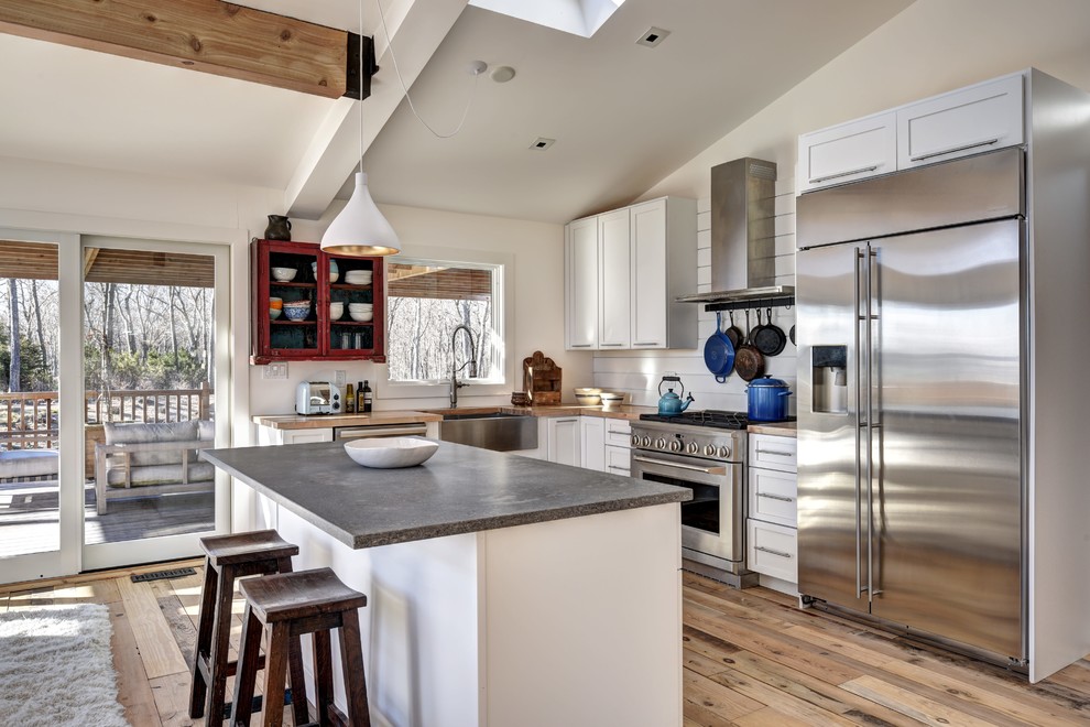 Example of a mid-sized mountain style l-shaped light wood floor and beige floor kitchen design in New York with a farmhouse sink, shaker cabinets, white cabinets, wood countertops, white backsplash, wood backsplash, stainless steel appliances, an island and beige countertops