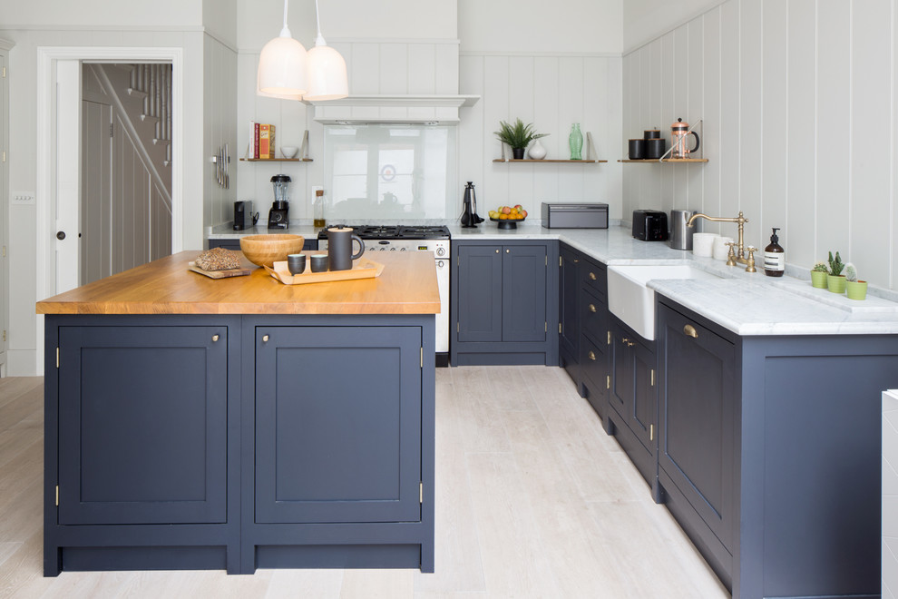 Bild på ett mellanstort vintage vit vitt l-kök, med en rustik diskho, blå skåp, marmorbänkskiva, grått stänkskydd, glaspanel som stänkskydd, svarta vitvaror, ljust trägolv, en köksö och skåp i shakerstil