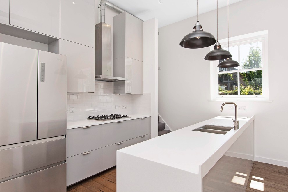 This is an example of a contemporary kitchen in London with a double-bowl sink, flat-panel cabinets, grey cabinets, white splashback, metro tiled splashback, stainless steel appliances, medium hardwood flooring, a breakfast bar and white worktops.