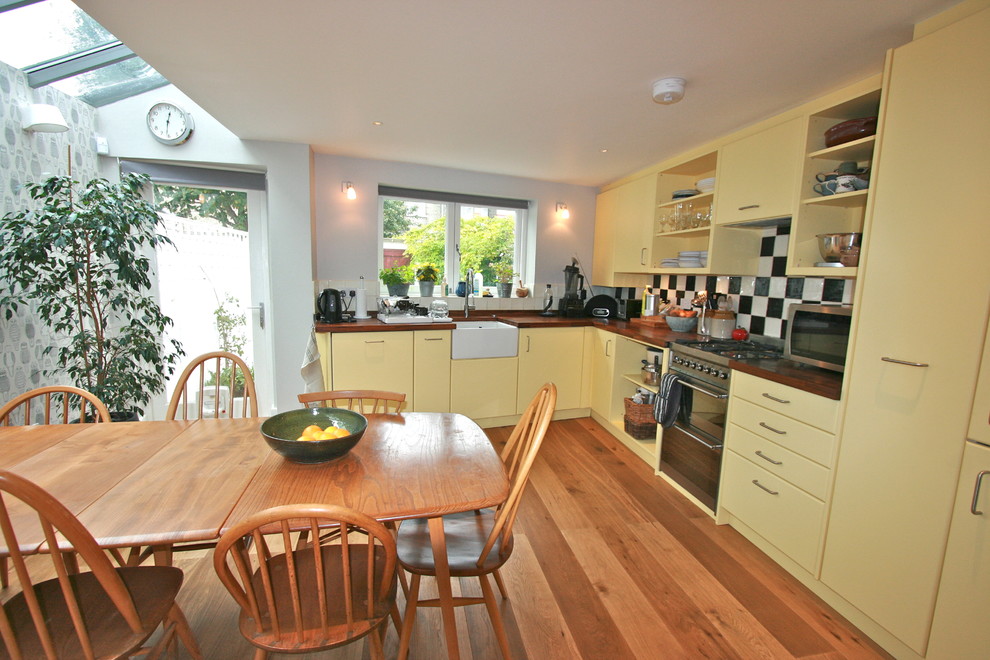Example of a mid-sized 1960s l-shaped medium tone wood floor eat-in kitchen design in London with a farmhouse sink, flat-panel cabinets, yellow cabinets, wood countertops, ceramic backsplash, stainless steel appliances and no island