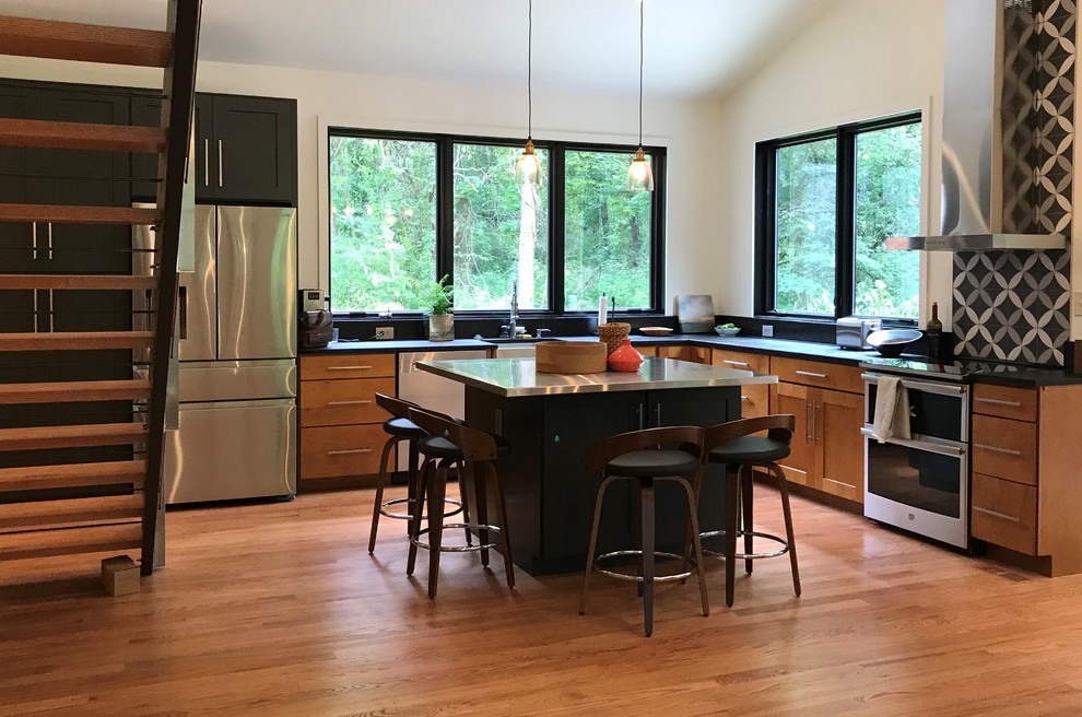 Photo of a medium sized contemporary l-shaped open plan kitchen in DC Metro with a belfast sink, flat-panel cabinets, medium wood cabinets, stainless steel worktops, black splashback, porcelain splashback, stainless steel appliances, medium hardwood flooring, an island, brown floors and black worktops.