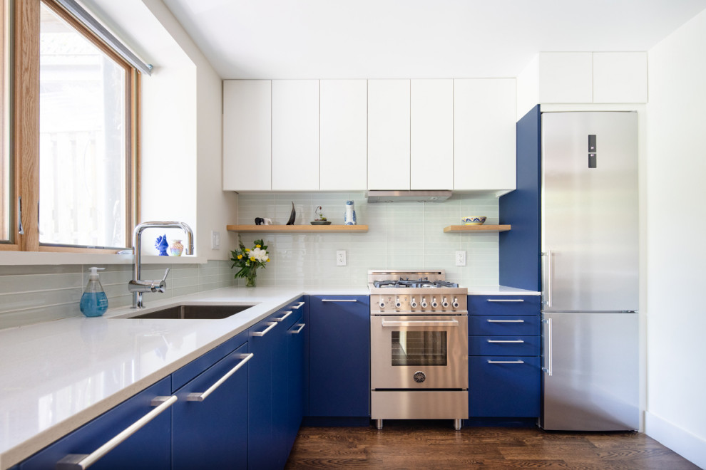 Moderne Küche in L-Form mit flächenbündigen Schrankfronten, blauen Schränken, Quarzwerkstein-Arbeitsplatte, weißer Arbeitsplatte, Unterbauwaschbecken, Küchenrückwand in Weiß, Rückwand aus Glasfliesen, dunklem Holzboden, braunem Boden und Küchengeräten aus Edelstahl in New York