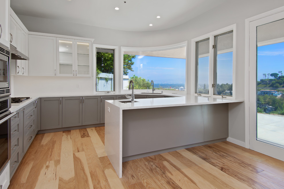 Photo of a medium sized classic u-shaped enclosed kitchen in Los Angeles with a submerged sink, shaker cabinets, grey cabinets, engineered stone countertops, stainless steel appliances, medium hardwood flooring, a breakfast bar, brown floors and white worktops.