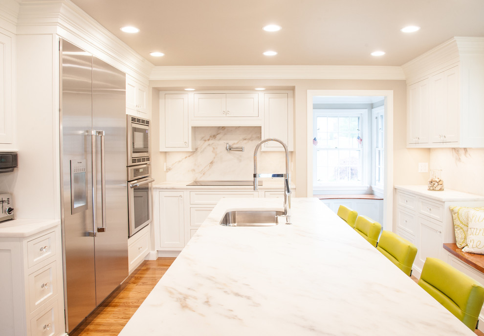 Klassisk inredning av ett kök, med en undermonterad diskho, luckor med profilerade fronter, vita skåp, marmorbänkskiva, vitt stänkskydd, stänkskydd i marmor, rostfria vitvaror och en köksö