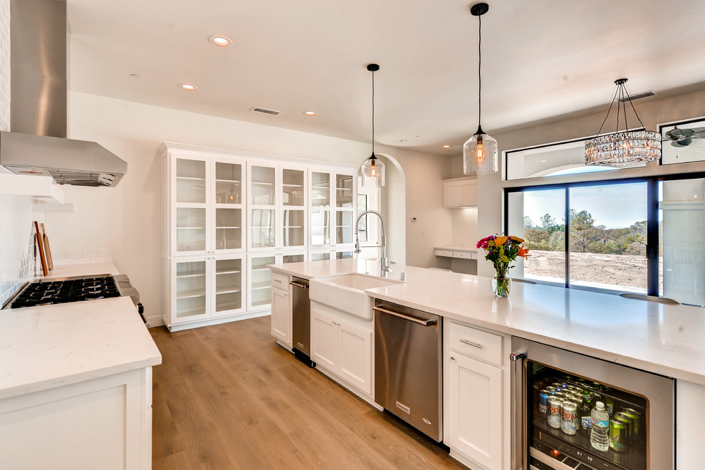 Example of a large farmhouse single-wall medium tone wood floor and brown floor eat-in kitchen design in Sacramento with a farmhouse sink, shaker cabinets, white cabinets, marble countertops, white backsplash, subway tile backsplash, stainless steel appliances, an island and white countertops