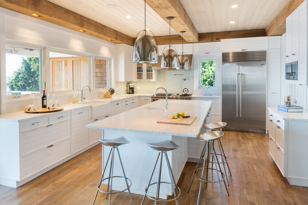 Photo of a large beach style l-shaped kitchen/diner in New York with a belfast sink, white cabinets, stainless steel appliances, medium hardwood flooring, an island, shaker cabinets and composite countertops.