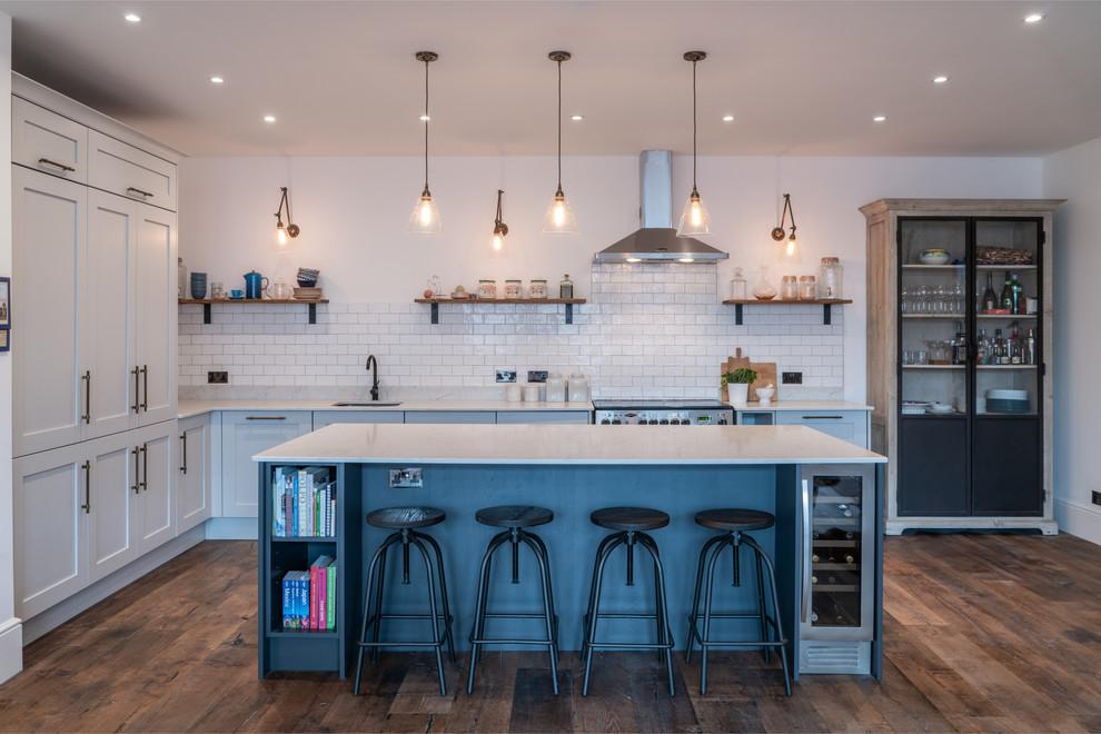 This is an example of a traditional l-shaped kitchen in London with a submerged sink, shaker cabinets, white cabinets, white splashback, metro tiled splashback, integrated appliances, medium hardwood flooring, an island, brown floors and white worktops.