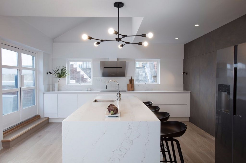 Inspiration for a contemporary kitchen in London with a submerged sink, flat-panel cabinets, white cabinets, white splashback, stone slab splashback, stainless steel appliances, light hardwood flooring, an island and white worktops.