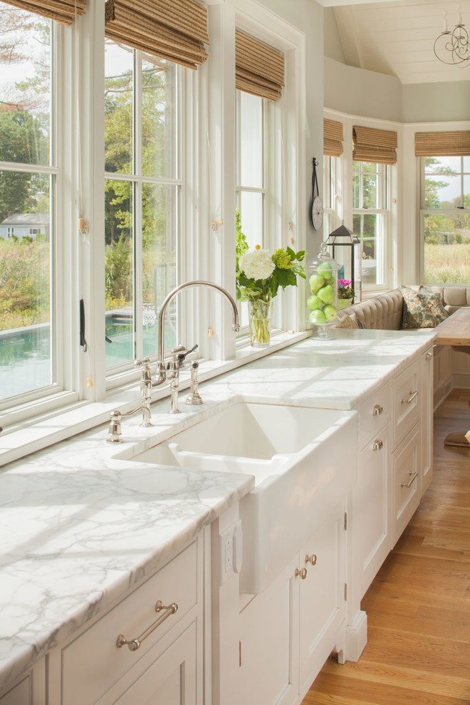 Example of a large beach style light wood floor eat-in kitchen design in New York with a farmhouse sink, recessed-panel cabinets, white cabinets, marble countertops, white countertops and window backsplash