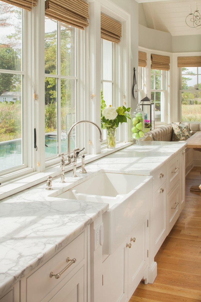 Example of a large beach style light wood floor eat-in kitchen design in New York with a farmhouse sink, recessed-panel cabinets, white cabinets, marble countertops and white countertops