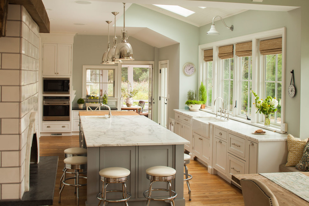 Example of a large beach style l-shaped light wood floor eat-in kitchen design in New York with a farmhouse sink, recessed-panel cabinets, white cabinets, marble countertops, white backsplash, stainless steel appliances and an island