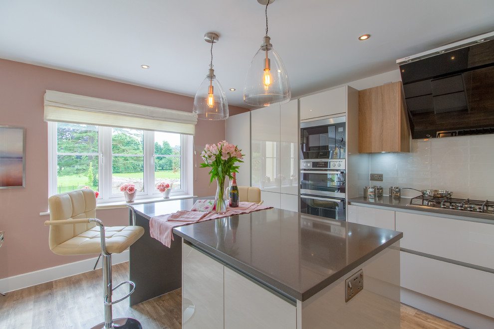 Einzeilige, Mittelgroße Moderne Küche mit flächenbündigen Schrankfronten, Küchengeräten aus Edelstahl, hellem Holzboden, Kücheninsel, weißen Schränken, Küchenrückwand in Weiß, braunem Boden und grauer Arbeitsplatte in Gloucestershire