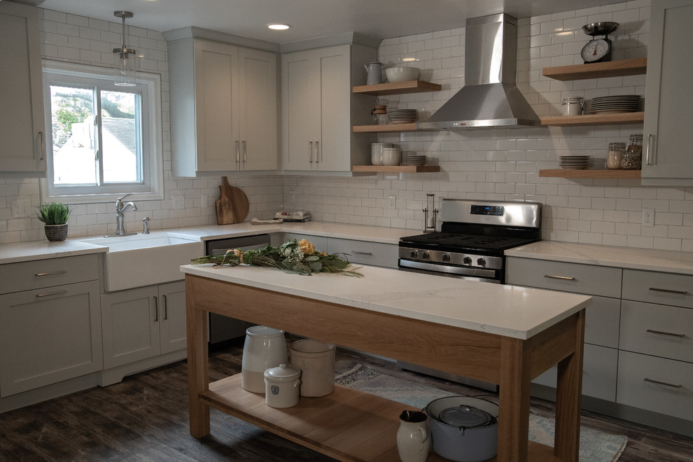 Inspiration for a mid-sized farmhouse l-shaped medium tone wood floor and gray floor enclosed kitchen remodel in Detroit with a farmhouse sink, recessed-panel cabinets, gray cabinets, quartz countertops, white backsplash, ceramic backsplash, stainless steel appliances, an island and white countertops