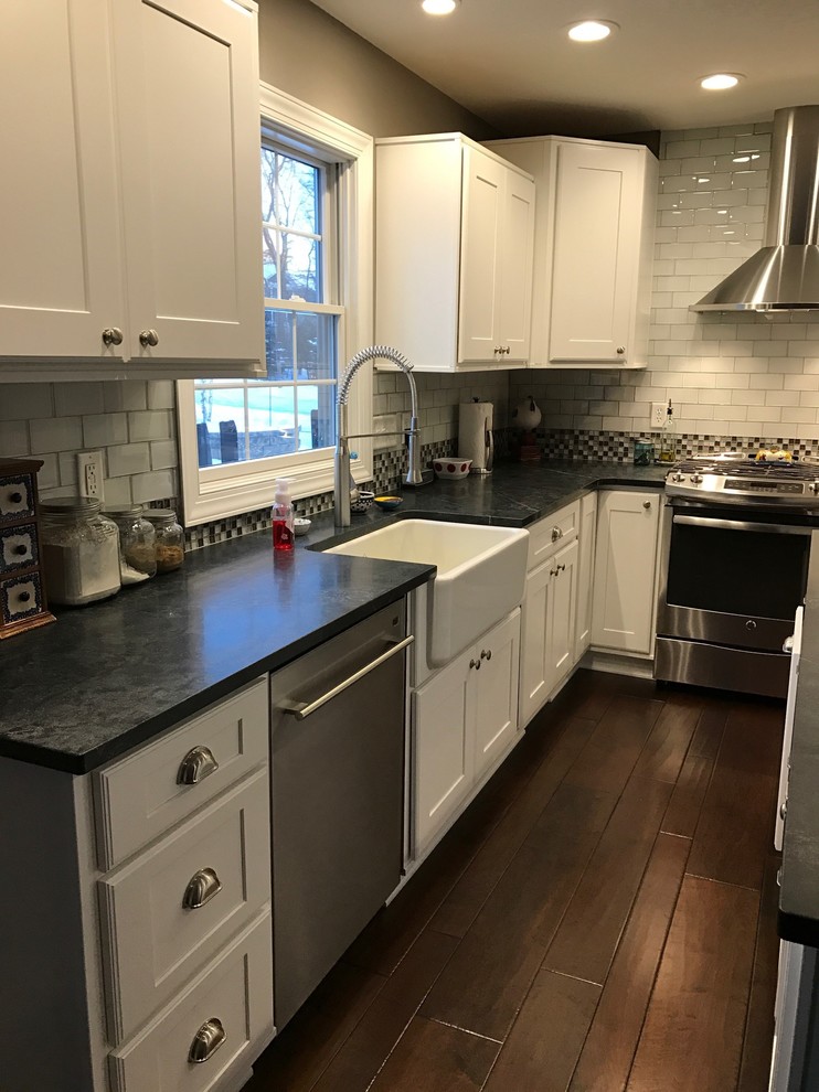 Inspiration for a mid-sized contemporary l-shaped medium tone wood floor and brown floor eat-in kitchen remodel in Cedar Rapids with a farmhouse sink, flat-panel cabinets, white cabinets, soapstone countertops, white backsplash, glass tile backsplash, stainless steel appliances, an island and gray countertops