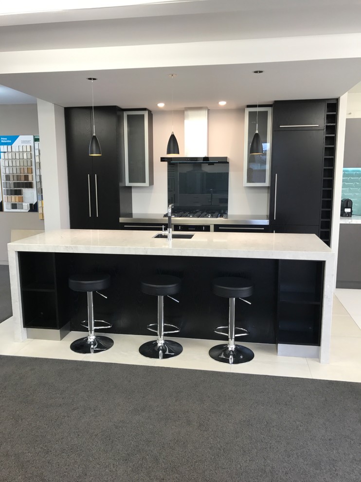 Photo of a medium sized contemporary galley open plan kitchen in Christchurch with a submerged sink, flat-panel cabinets, black cabinets, engineered stone countertops, black splashback, glass sheet splashback, stainless steel appliances, ceramic flooring, an island and white floors.