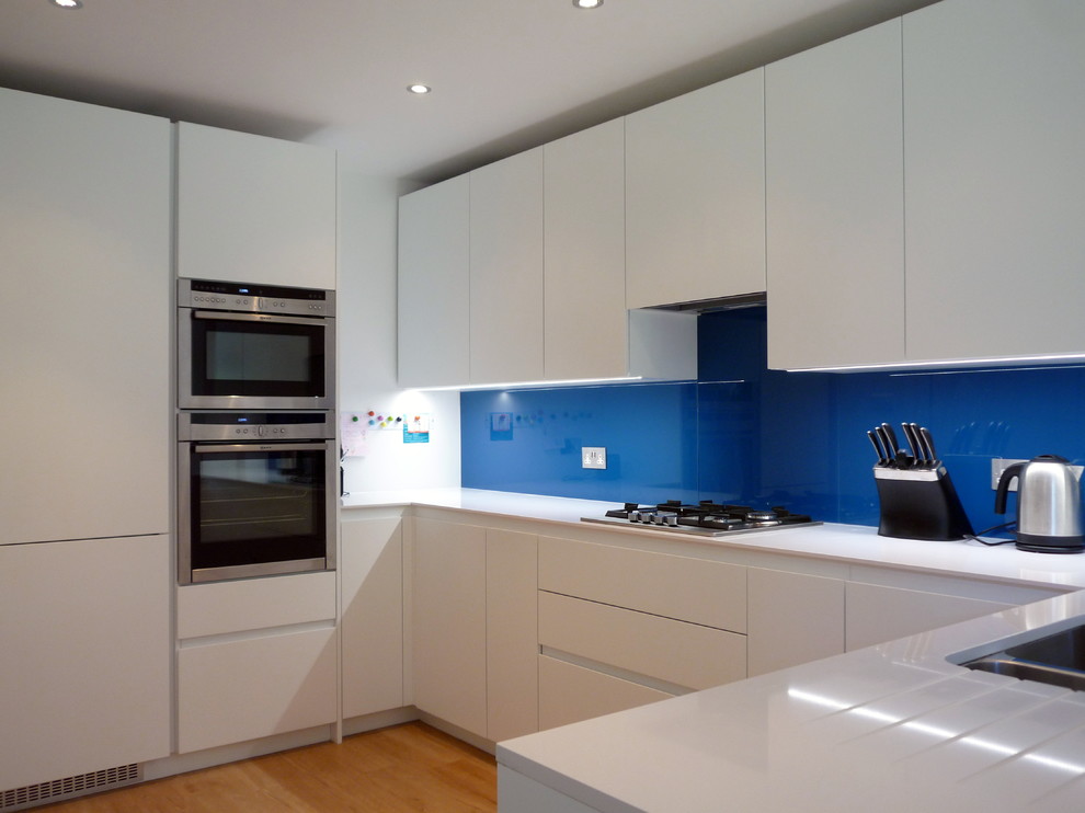 Inspiration for a medium sized modern u-shaped kitchen/diner in London with a submerged sink, flat-panel cabinets, white cabinets, engineered stone countertops, blue splashback, glass sheet splashback, stainless steel appliances, light hardwood flooring and no island.