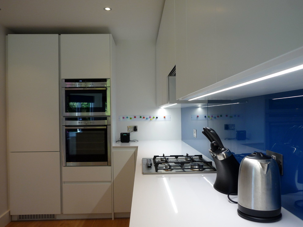 This is an example of a medium sized modern u-shaped kitchen/diner in London with a submerged sink, flat-panel cabinets, white cabinets, engineered stone countertops, blue splashback, glass sheet splashback, stainless steel appliances, no island and light hardwood flooring.