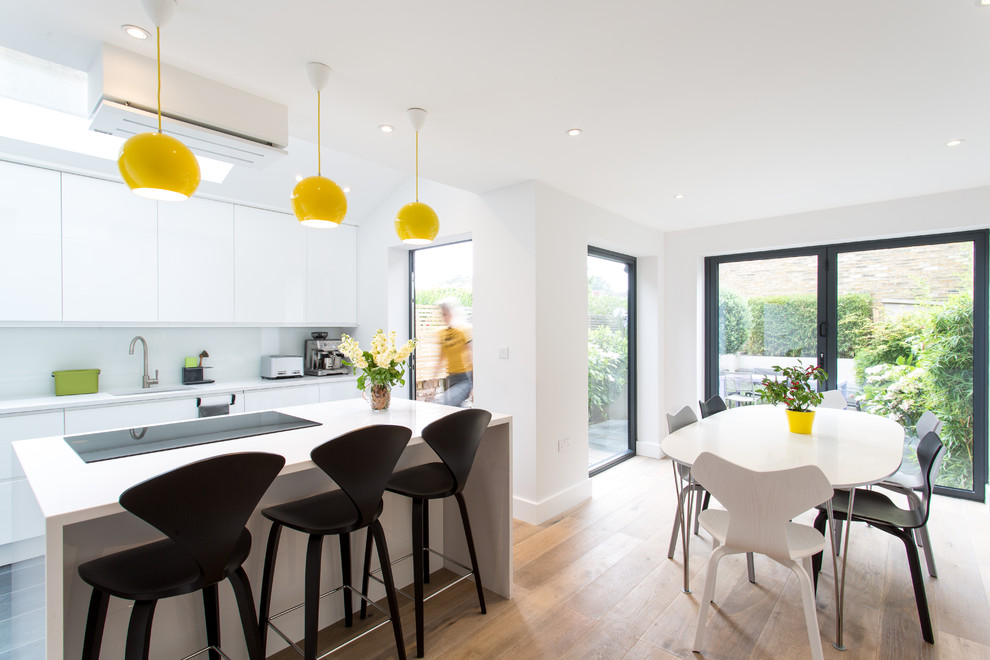 This is an example of a medium sized contemporary single-wall kitchen/diner in Other with an integrated sink, flat-panel cabinets, white cabinets, composite countertops, white splashback, glass sheet splashback, integrated appliances, light hardwood flooring and an island.