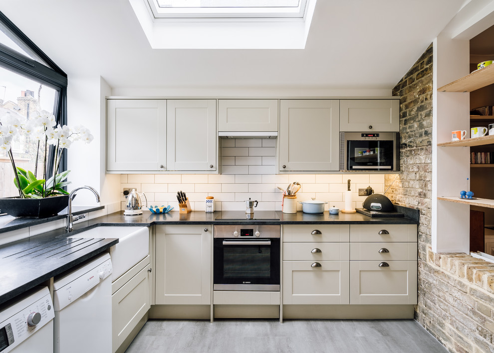 Photo of a classic l-shaped kitchen in London with a belfast sink, shaker cabinets, beige cabinets, white splashback, stainless steel appliances, medium hardwood flooring, grey floors and black worktops.