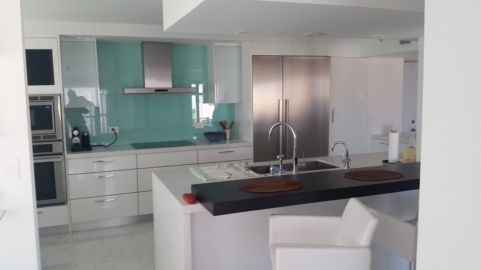 Photo of a large modern u-shaped kitchen/diner in Miami with a submerged sink, flat-panel cabinets, white cabinets, quartz worktops, green splashback, glass sheet splashback, stainless steel appliances, porcelain flooring and multiple islands.