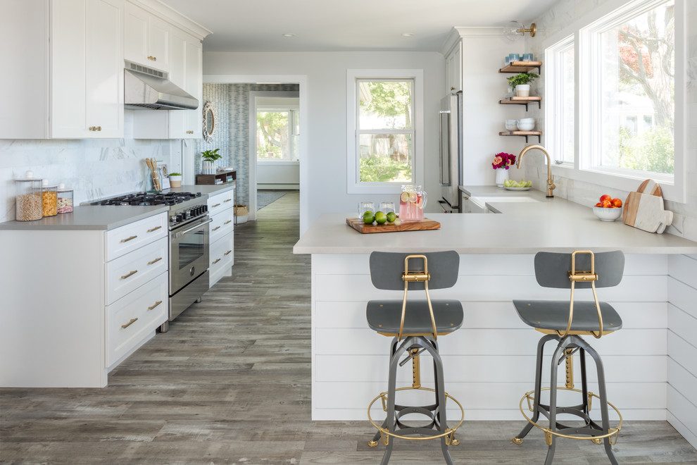 Example of a beach style light wood floor enclosed kitchen design in New York with a farmhouse sink, shaker cabinets, white cabinets, white backsplash, marble backsplash, stainless steel appliances, a peninsula and gray countertops