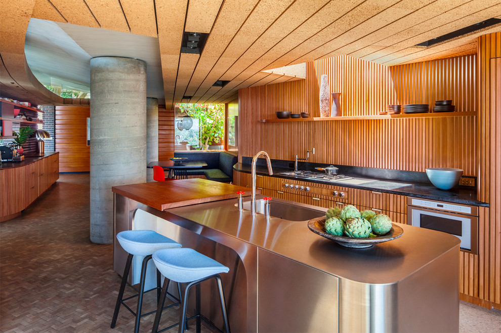 Mid-Century Wohnküche mit integriertem Waschbecken, hellbraunen Holzschränken, Edelstahl-Arbeitsplatte, Küchenrückwand in Braun, Rückwand aus Holz, Elektrogeräten mit Frontblende, Kücheninsel und braunem Boden in Los Angeles