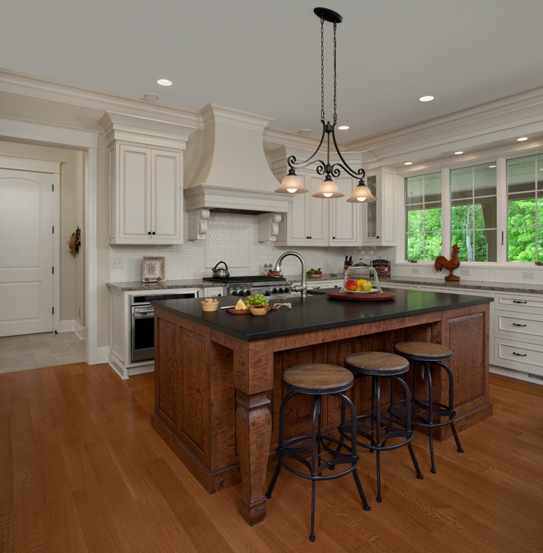 Eat-in kitchen - huge traditional l-shaped medium tone wood floor and brown floor eat-in kitchen idea in Grand Rapids with a farmhouse sink, flat-panel cabinets, white cabinets, granite countertops, white backsplash, ceramic backsplash, stainless steel appliances and an island