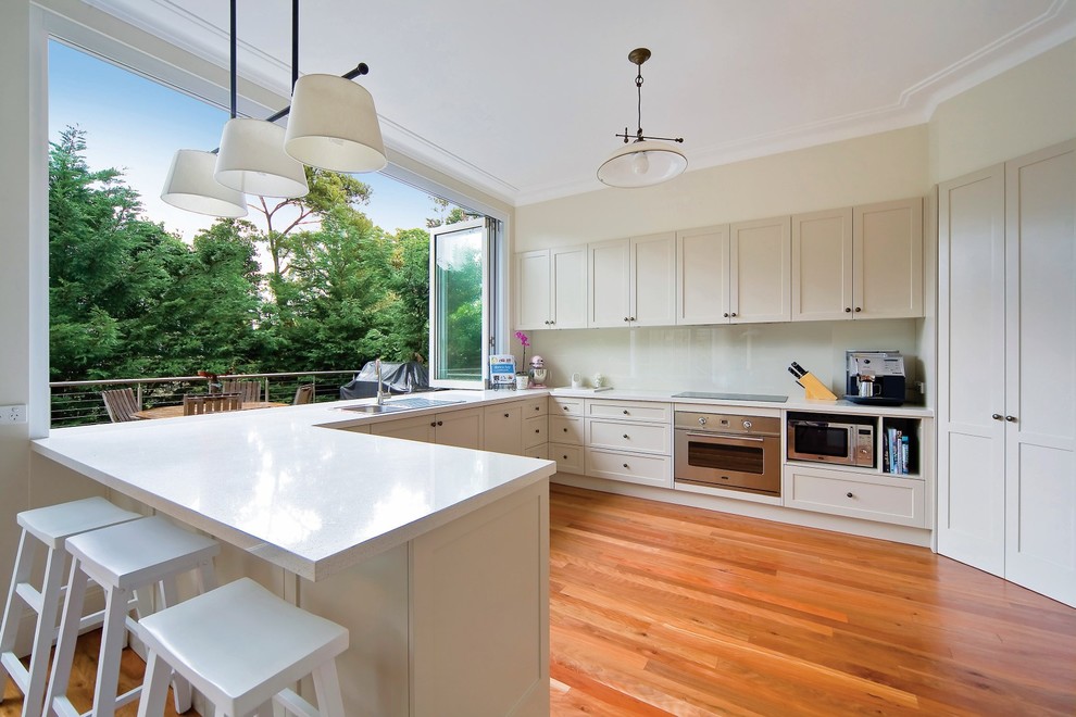 This is an example of a classic u-shaped kitchen/diner in Sydney with white cabinets, white splashback, stainless steel appliances, medium hardwood flooring, white worktops, a built-in sink, shaker cabinets, glass sheet splashback, a breakfast bar and red floors.