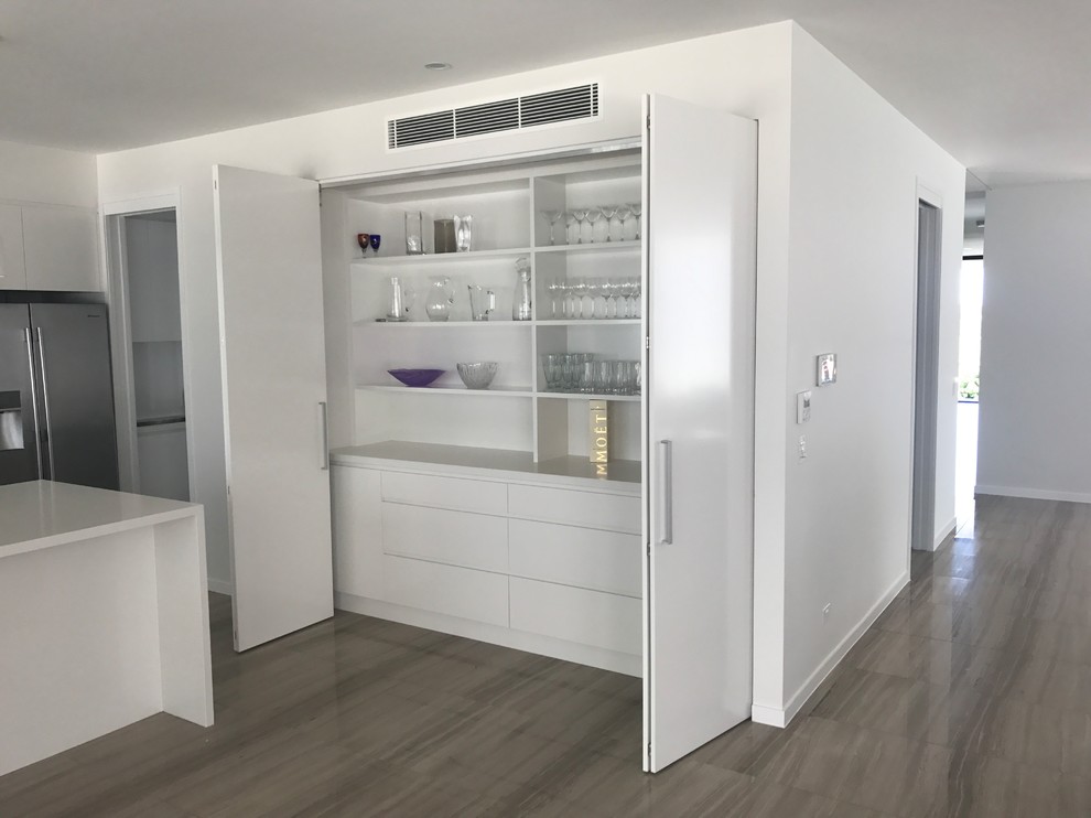 Large modern l-shaped kitchen pantry in Brisbane with a submerged sink, flat-panel cabinets, white cabinets, engineered stone countertops, white splashback, glass sheet splashback, stainless steel appliances, marble flooring, an island and brown floors.