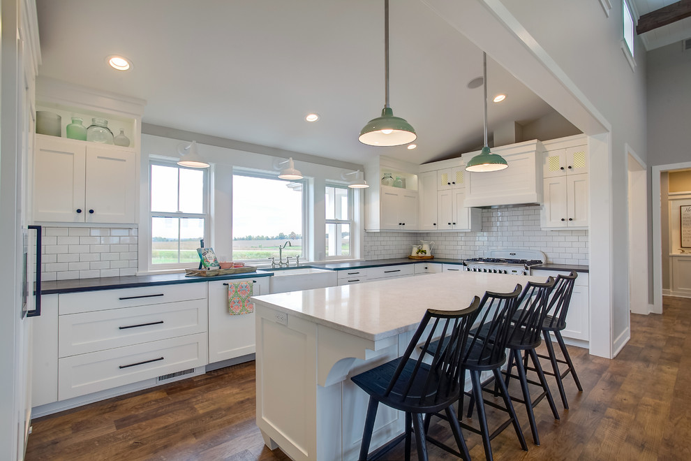 Photo of a rural u-shaped kitchen in Grand Rapids with a belfast sink, shaker cabinets, white cabinets, white splashback, metro tiled splashback, integrated appliances, medium hardwood flooring, an island, brown floors and white worktops.
