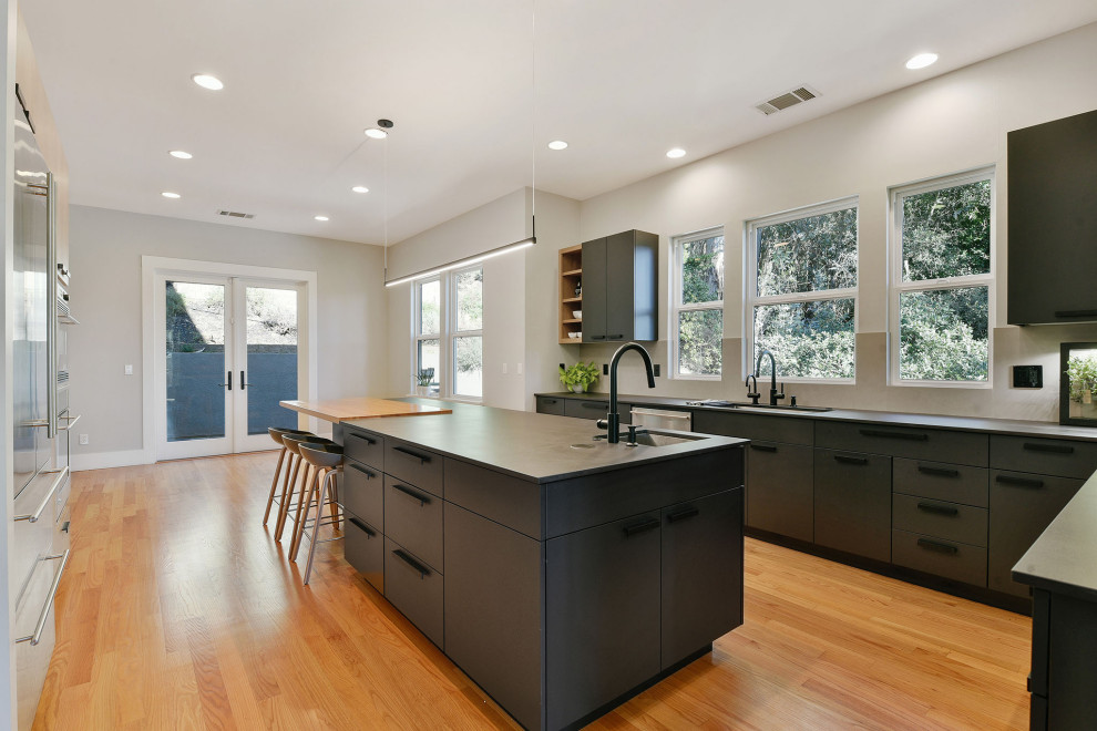 Inspiration for a large modern enclosed kitchen in San Francisco with a double-bowl sink, flat-panel cabinets, grey splashback, stone slab splashback, stainless steel appliances, light hardwood flooring, an island and black worktops.