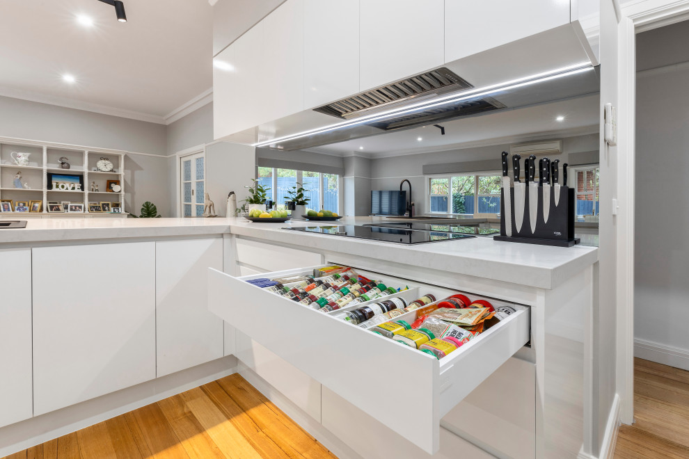 This is an example of a large modern l-shaped open plan kitchen in Melbourne with a double-bowl sink, flat-panel cabinets, white cabinets, engineered stone countertops, mirror splashback, black appliances and light hardwood flooring.