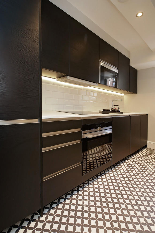 Enclosed kitchen - small contemporary galley ceramic tile enclosed kitchen idea in London with a single-bowl sink, flat-panel cabinets, dark wood cabinets, solid surface countertops, white backsplash, porcelain backsplash, black appliances and no island