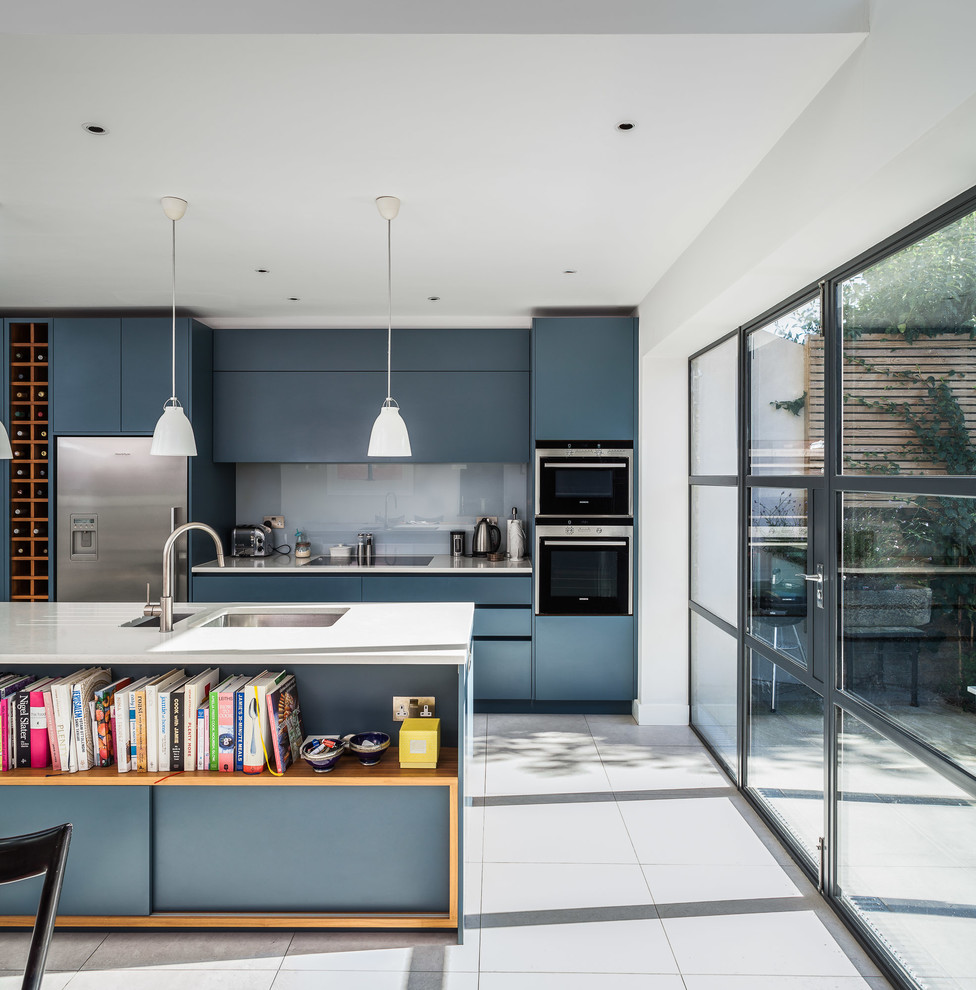 Стильный дизайн: параллельная кухня в современном стиле с врезной мойкой, плоскими фасадами, синими фасадами, белым фартуком, фартуком из стекла, техникой из нержавеющей стали и островом - последний тренд