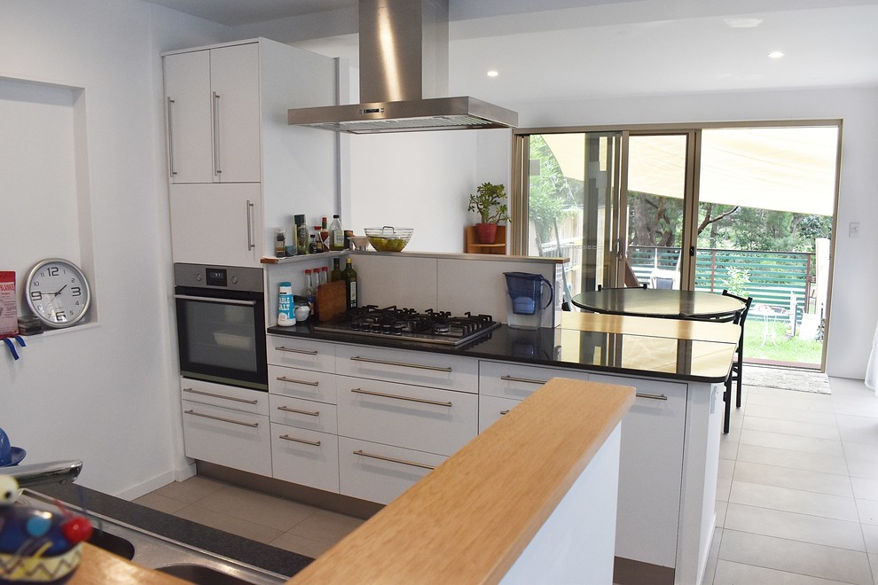 Photo of a small contemporary galley kitchen/diner in Gold Coast - Tweed with white cabinets, granite worktops, ceramic flooring, an island, grey floors, black worktops, a built-in sink, open cabinets, white splashback, wood splashback and black appliances.