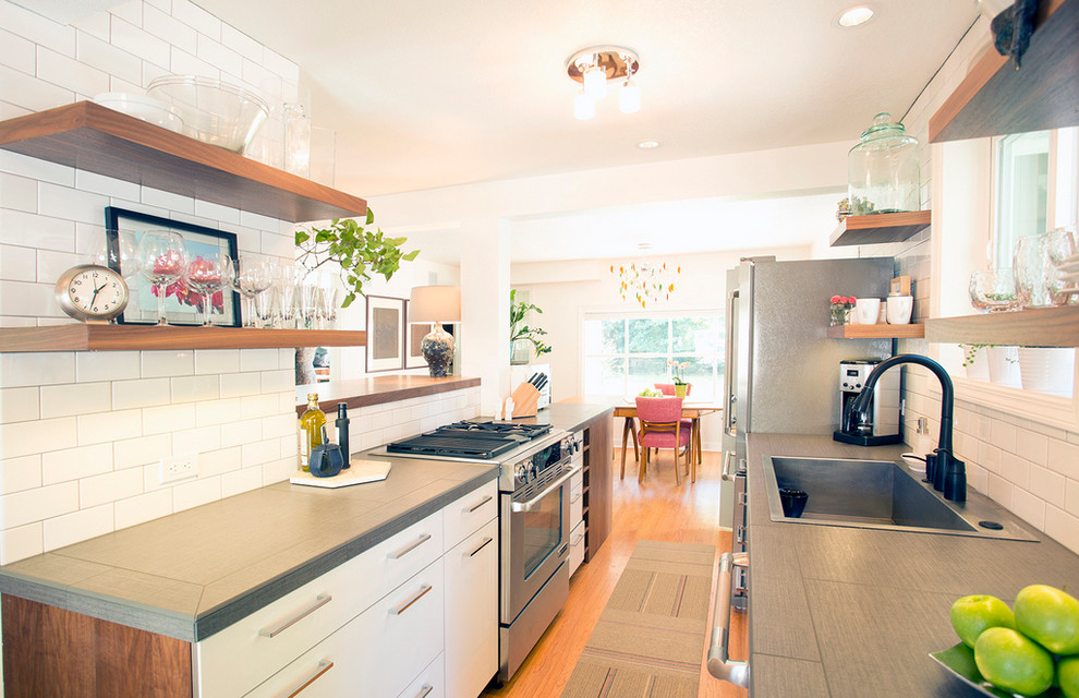 Example of a small transitional galley light wood floor open concept kitchen design in Portland with a drop-in sink, flat-panel cabinets, white cabinets, tile countertops, white backsplash, subway tile backsplash, stainless steel appliances and no island