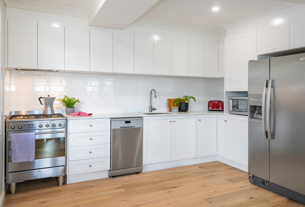 This is an example of a small traditional l-shaped open plan kitchen in Brisbane with a submerged sink, shaker cabinets, white cabinets, engineered stone countertops, white splashback, metro tiled splashback, stainless steel appliances, light hardwood flooring, no island and white worktops.