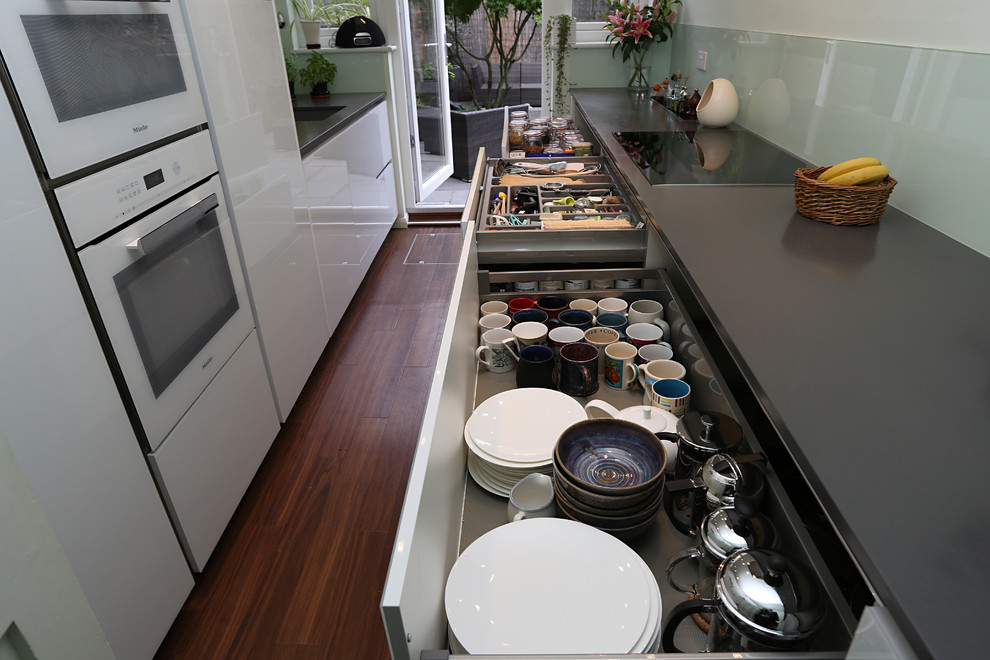 Geschlossene, Zweizeilige, Mittelgroße Moderne Küche ohne Insel mit Waschbecken, Glasfronten, weißen Schränken, Quarzit-Arbeitsplatte, Glasrückwand, weißen Elektrogeräten und braunem Holzboden in London