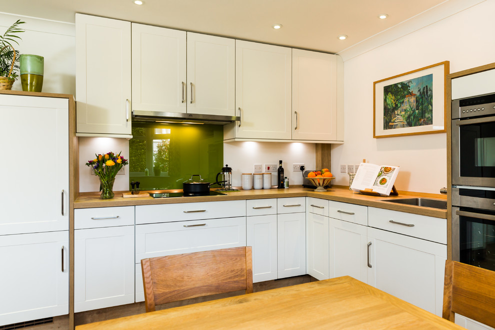 This is an example of a small classic l-shaped kitchen/diner in Hampshire with a single-bowl sink, shaker cabinets, white cabinets, laminate countertops, green splashback, glass sheet splashback, stainless steel appliances and light hardwood flooring.