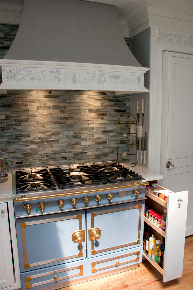 Bild på ett avskilt, mellanstort vintage l-kök, med en undermonterad diskho, luckor med profilerade fronter, vita skåp, bänkskiva i återvunnet glas, blått stänkskydd, stänkskydd i glaskakel, integrerade vitvaror, ljust trägolv och en köksö